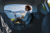 Fototapeta Koty - Young woman sitting in the open trunk of a car overlooking the sea and using a laptop, freelancer, remote work and summer vacation auto travel