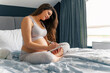 © DusanJelicic - A pregnant woman with long brown hair is sitting on the bed in front of the window with her legs crossed as she writes something on her tablet