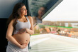 © DusanJelicic - a pregnant woman with long brown hair stands leaning against the window with her hands gently on her stomach and looking out the window
