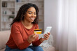 © Prostock-studio - Smiling black woman at home holding credit card and smartphone, shopping online
