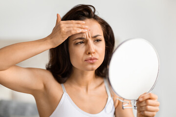 Upset millennial woman looking at mirror, checking pimple