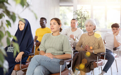 Canvas Print - Positive young woman visiting seminar during educational classes, listening carefully to lecturer with multiethnic group of coursemates on different ages..