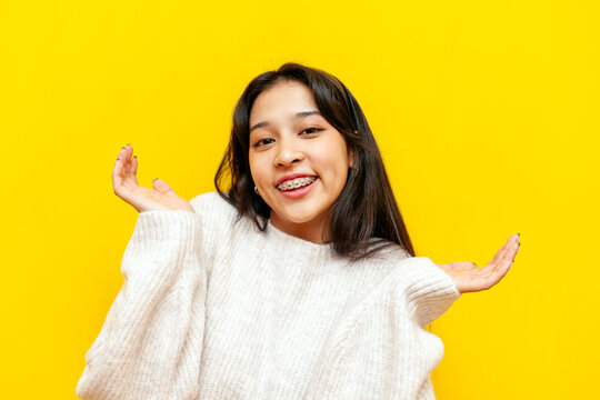 young unsure asian woman with braces shrugs over yellow isolated background, korean girl doesn't know the answer and is uncertain and confused