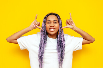 Wall Mural - young african american woman with hipster colored dreadlocks smiling and showing youth gesture with hands on yellow isolated background, girl with unique purple hairstyle looking at camera