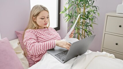 Sticker - A young woman works on a laptop while lying on a bed in her bedroom, with papers and a serene decor.