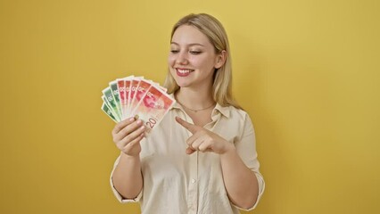 Sticker - Cheerful young blonde woman flashing a confident smile as she points at israeli shekel banknotes, creating a vibrant and hopeful scene against an isolated yellow background.