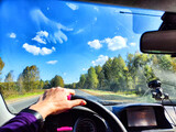 Fototapeta Tulipany - view from a car windshield of natural landscape with road, green trees and blue sky in summer or spring time. Hand of woman on the steering wheel. Female traveler driving alone on trip or journey