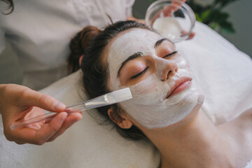 Wall Mural - Professional cosmetologist applying moisturizing mask on face of a woman. Attractive young lady getting skin care treatmen.