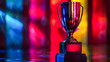 A trophy is sitting on a table in front of a colorful background. The trophy is shiny and metallic, and it is placed on a black base. The colorful background adds a sense of excitement