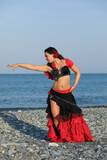Fototapeta Perspektywa 3d - Dancer woman in black and red suit with fan dancing on seashore, gesture