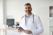 © Home-stock - Professional help. Happy middle aged male general practitioner standing with tablet and smiling at his office in clinic