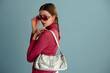 Fashionable confident woman wearing pink wraparound sunglasses, turtleneck, white skirt, with trendy metallic leather bag posing on blue background. Studio fashion portrait. Copy, empty space for text