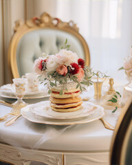 Still life of a stack of pancakes with flowers on top in front of a blurry background of a table set with fine china.