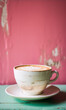 Pink background, white coffee cup with latte art on a saucer on a green table.