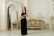 Pretty woman in black dress with sax poses in baroque studio with sofa, mirror