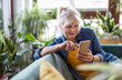 © pikselstock - Smiling senior woman using smart phone while sitting on sofa at home