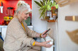 © pikselstock - Senior woman holding mobile phone while adjusting the thermostat on the radiator at home
