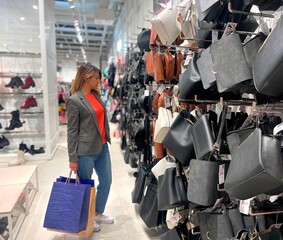 Side view of Caucasian pretty attractive woman standing in store indoor looking at bags at shopping. Adult young female buying bag in city mall. Buyer concept. Season sale