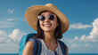 Cheerful Asian tourist woman, accessorized with a beach hat, sunglasses, and backpack, ready to embark on holiday escapades with a backdrop of azure skies.