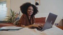 Portrait of cheerful self-employed woman with curly hair working on laptop in relaxed pose in bed