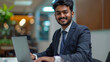 © Muhammad - A smiling young Indian man in a suit sits in the office at a desk and works on a laptop, typing on a keyboard, texting, working with data