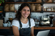 © Sascha - Portrait of smiling Hispanic woman in a cafe, working on a laptop at a counter, wearing denim overall and a white t-shirt, small business concept.