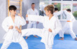 Focused teenage boys in white kimonos practicing karate kicking techniques during sparring under supervision of enthusiastic coach..