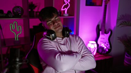 Canvas Print - Cheeky young hispanic man, a master streamer, confidently sporting headphones, smiling with crossed arms, chilling in his cozy gaming room at night, surrounded by technology.