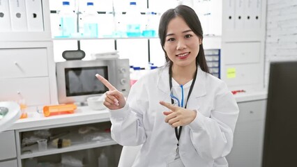 Canvas Print - Cheerful young chinese woman scientist playfully points to the side, confidently smiling at the camera. she's welcoming us into her buzzing lab, eager to share the joy of science!