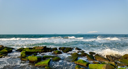 Wall Mural - Vibrant seascape with moss-covered rocks on the shore, white foamy waves crashing, and distant islands under a clear blue sky, ideal for background with space for text on the right