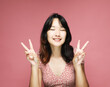 Young asian woman show victory sign , experiencing exciting emotions on pink background