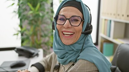 Sticker - A smiling mature woman wearing a hijab and headset working as a customer service representative in a modern office.