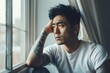 depressed asian american man in white t-shirt with tattoo on arms looking through window