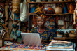 Creative African female shop owner multitasking in craft workshop: writing notes, using laptop.