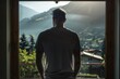Backlit silhouette of a man in a simple t-shirt, standing before a window with a panoramic view of a mountainous landscape. The silhouette reveals the contours of his tattooed arms.