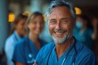 A portrait of a happy middle-aged male doctor in scrubs standing and smiling with his team behind him at the hospital, in a wide shot with soft light and natural colors