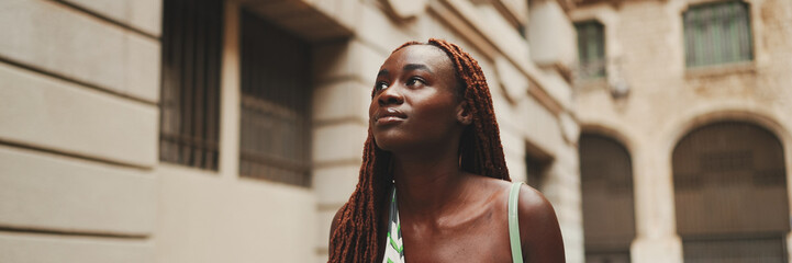 Wall Mural - Gorgeous woman with African braids dressed in top walks down the street, Panorama. Stylish smiling girl looks at the upper floors of buildings