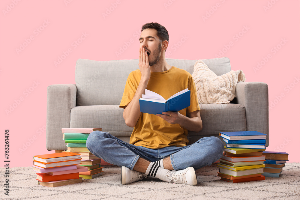 Sleepy young man with books sitting near sofa on pink background
