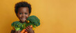 A studio shot of a smiling black boy holding fresh broccoli and carrots on a yellow background. Copying the space. The concept of healthy baby food.