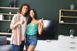 A diverse couple is posing with thumbs up in a cozy living room with copy space
