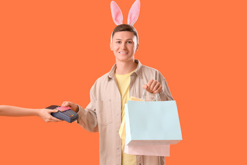 Sticker - Young man in bunny ears with shopping bags paying via terminal on orange background. Easter Sale