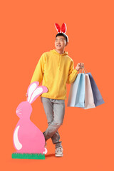 Wall Mural - Young Asian man in bunny ears with paper rabbit and shopping bags on orange background. Easter Sale