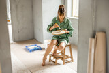 Fototapeta Przestrzenne - Young woman paints walls while making repairment of a new apartment. Sitting on chair and choosing paint color. Creative process of home renovation and repair concept