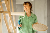 Fototapeta Do przedpokoju - Portrait of a young joyful and cute woman standing with paint roller during repairing process of a house. Concept of happy leisure time while renovating interior