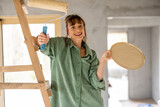 Fototapeta Sypialnia - Portrait of a young joyful and cute woman standing with paint roller during repairing process of a house. Concept of happy leisure time while renovating interior