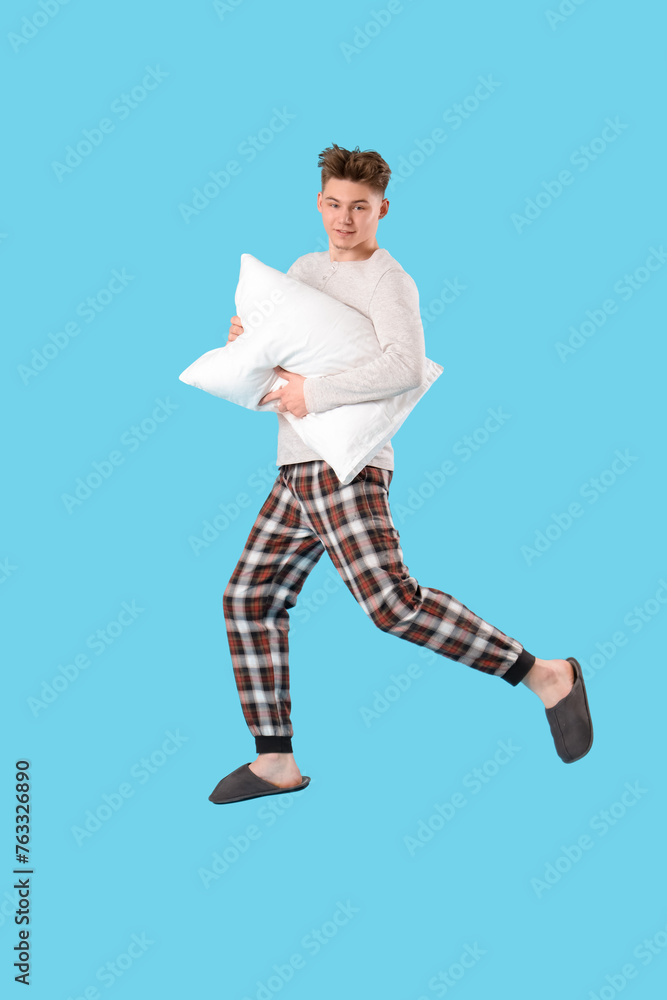 Jumping young man in pajamas with soft pillow on blue background