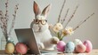Cute trendy stylish rabbit works on laptop surrounded flowers and Easter eggs. Bunny boss with glasses sitting at table. hipster style. Happy spring holiday, funny celebration. Humor Greeting card