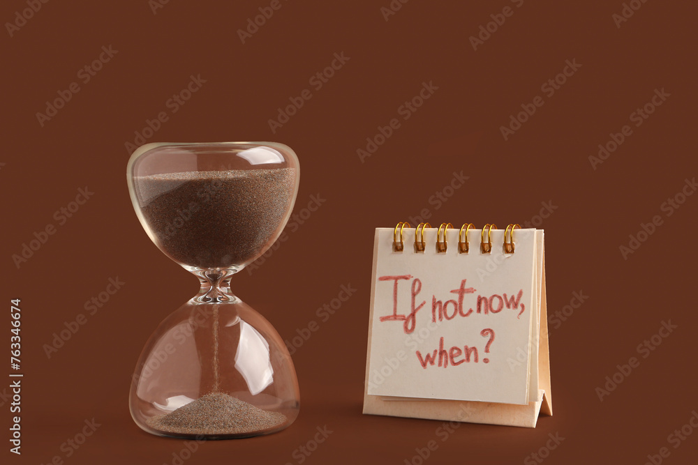 Notebook with text IF NOT NOW, WHEN? and hourglass on brown background