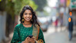 Portrait of a young Indian woman in a national green suit using a mobile phone while walking outside in the city, looking and smiling at the camera