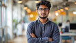 Portrait of a young successful Indian male programmer, developer standing in an office center, crossing his arms over his chest and looking seriously at the camera